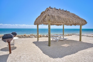 A beach with a thatched-roof gazebo, picnic tables, and barbecue grills. - Sandpiper Cove updated 2bed/2bath beachfront condo with shared pool (Marathon)