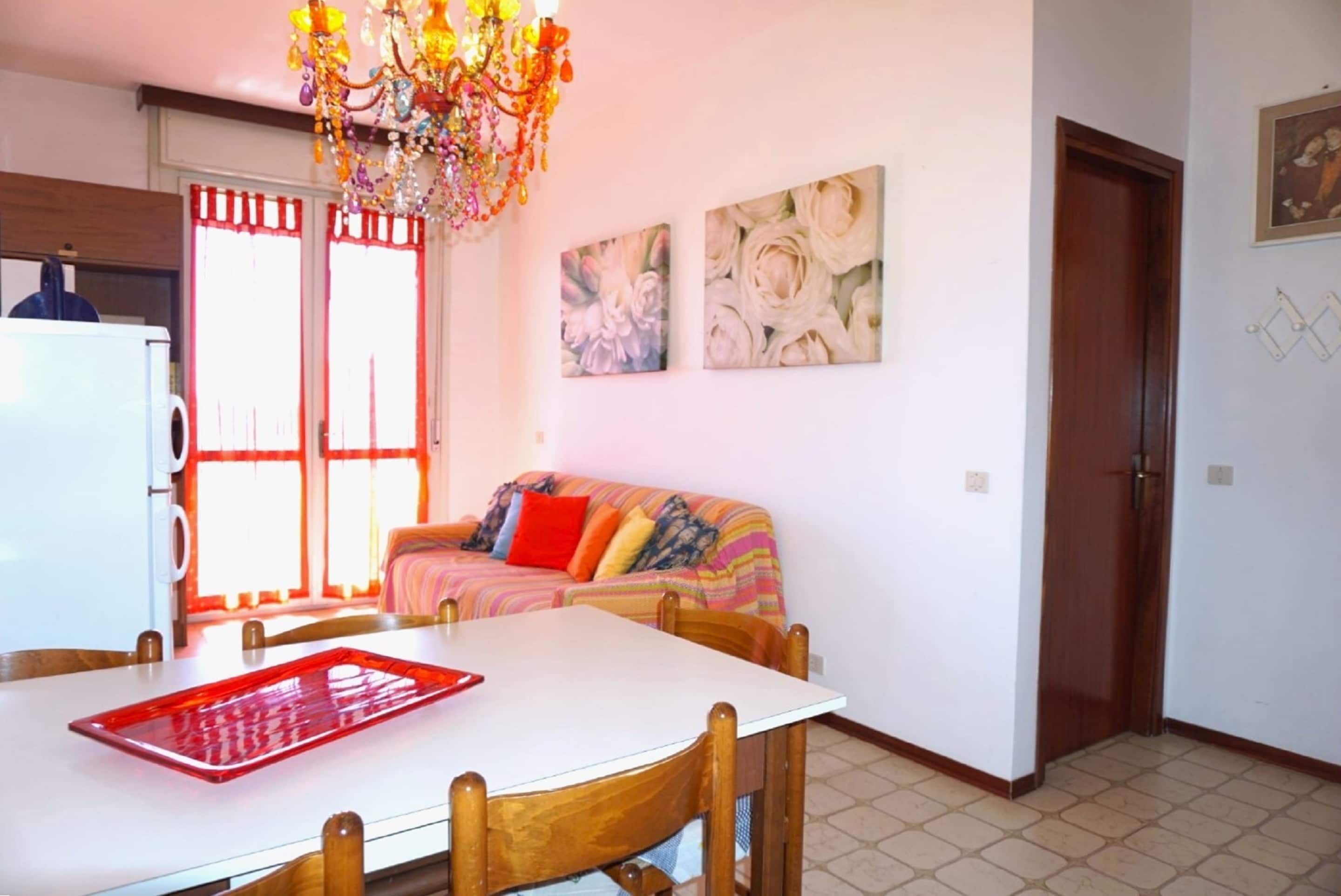 A dining area with a white table, wooden chairs, a chandelier, and a colorful sofa.