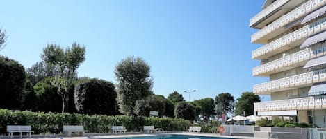 A swimming pool with a clear blue water surface, surrounded by a paved area with benches and a multi-story building with balconies in the background.