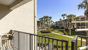 A balcony with a view of a courtyard featuring palm trees, a pathway, and a building with balconies. - Oceanview townhouse style condo in the heart of St. Augustine Beach! (Saint Augustine)