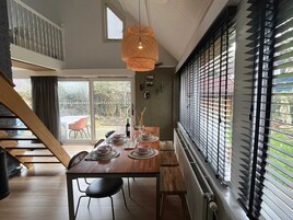 Un comedor con una mesa de madera dispuesta para cuatro personas, una lámpara colgante y vistas a través de una ventana con persianas.