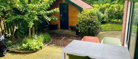 A patio with a table and chairs, surrounded by greenery and a wooden building with a blue roof.