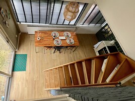 Un comedor con una mesa de madera dispuesta para cuatro personas, una escalera y vistas al exterior a través de amplios ventanales.