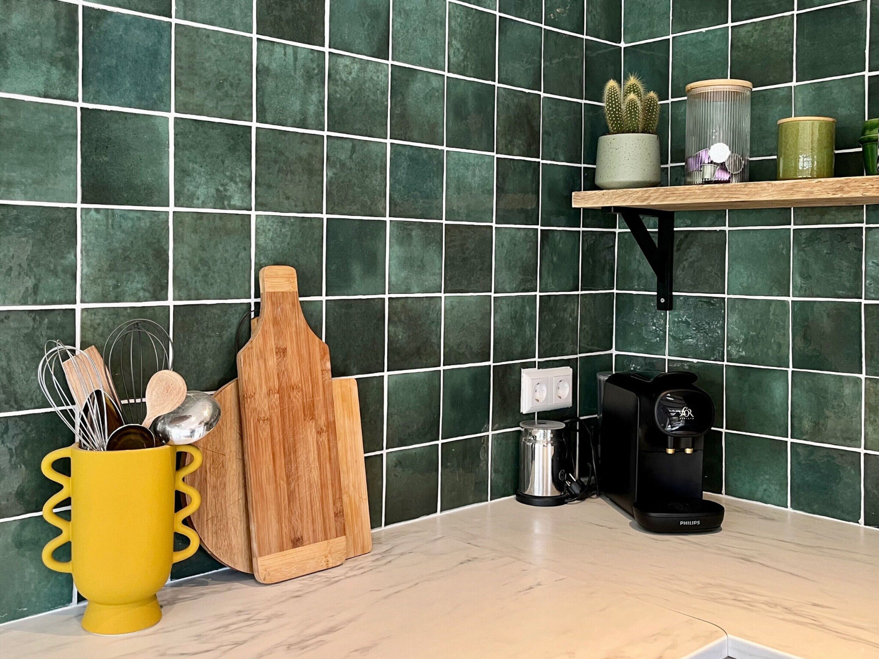 Ett kök med gröna kakelplattor ovanför arbetsbänken, ett gult förvaringsställ för köksredskap, en skärbräda i trä, en kaffebryggare och en hylla med en kaktus och glasburkar.