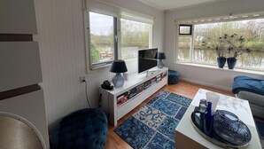 Un salon avec un téléviseur, une lampe, un pouf bleu, un tapis à motifs et une vue sur un lac par les fenêtres.