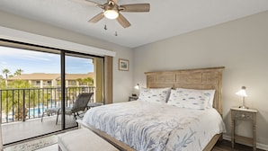Une chambre dotée d’un grand lit, d’un ventilateur de plafond, d’une table de chevet avec une lampe, et offrant une vue sur une piscine et des palmiers, grâce à une porte-fenêtre coulissante.