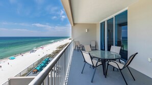 Un balcon avec une table et des chaises en verre, donnant sur une plage parsemée de parasols et fréquentée par des gens.