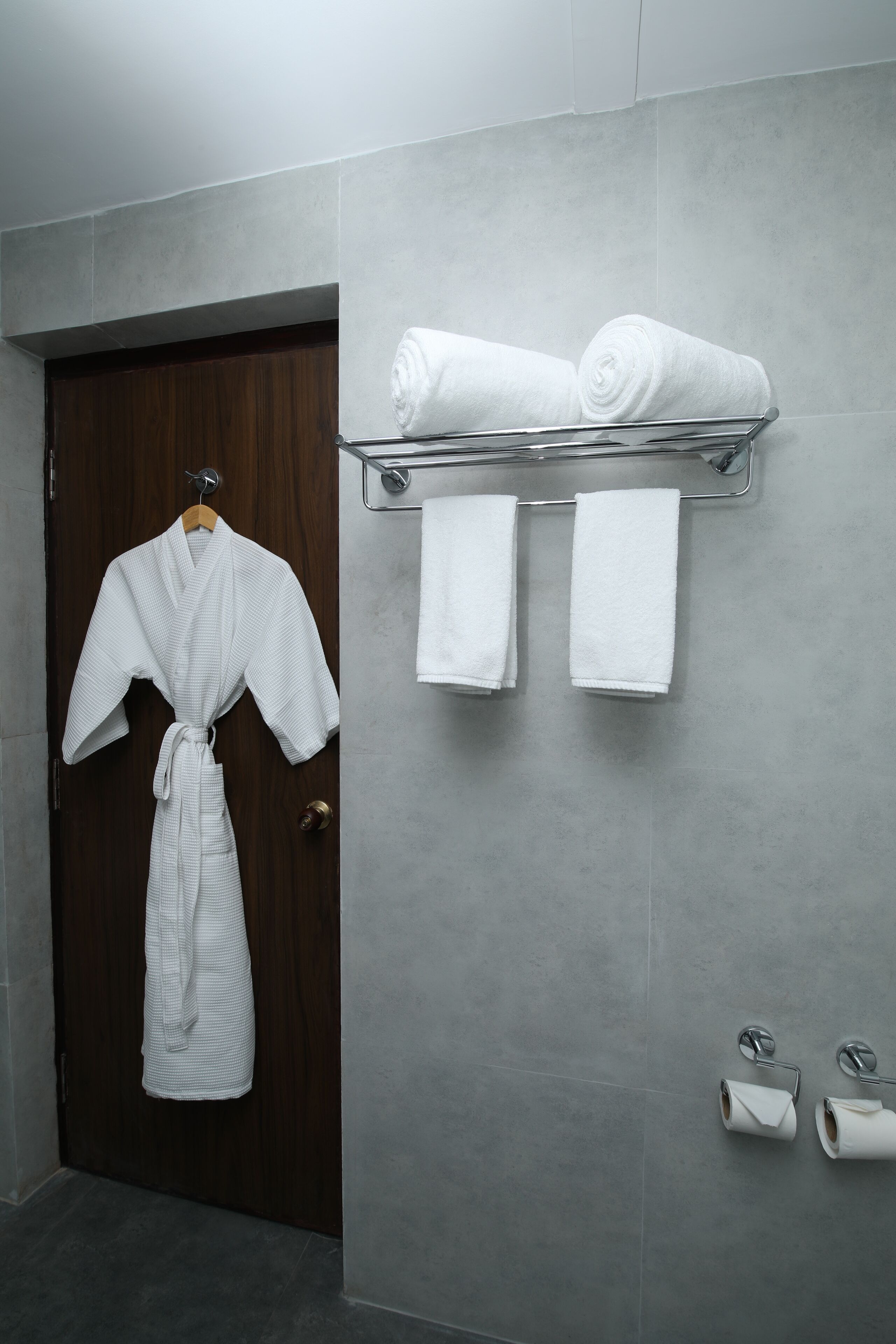 A bathroom with a wooden door, a metal towel rack holding two rolled towels, and two toilet paper rolls on the floor.