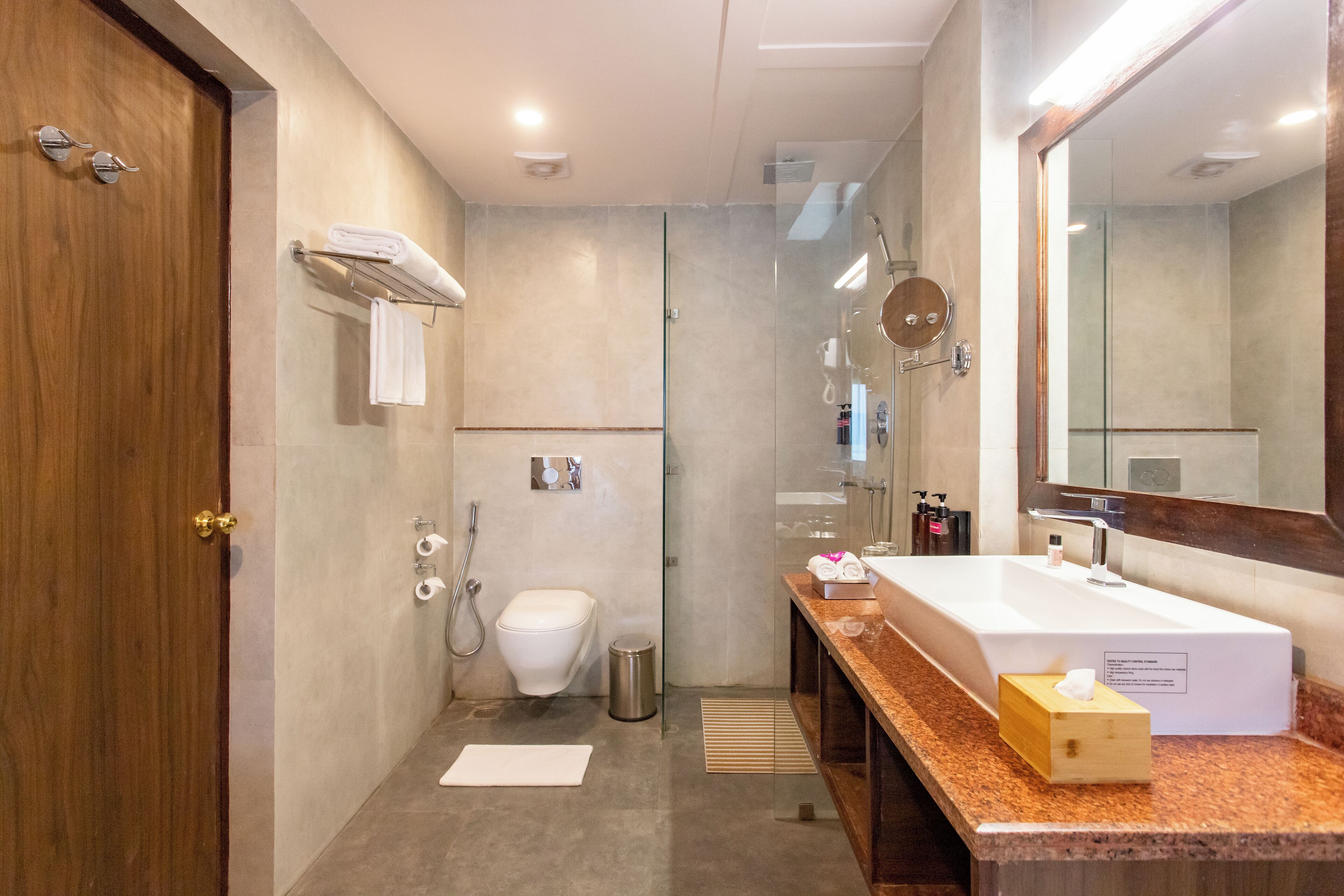 Un bagno moderno con porta in legno, un water bianco, una doccia vetrata e un piano lavabo in marmo con lavandino e articoli da toilette.