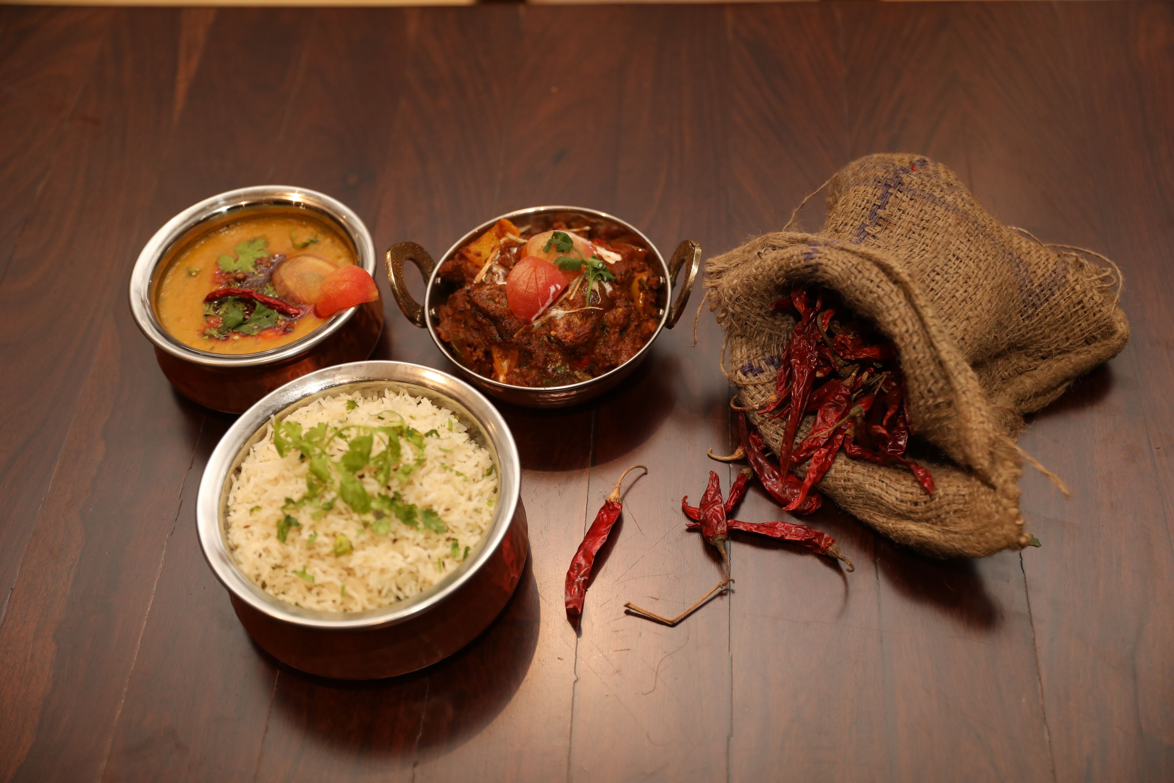 Three bowls of food and a sack of dried chilies on a wooden table.