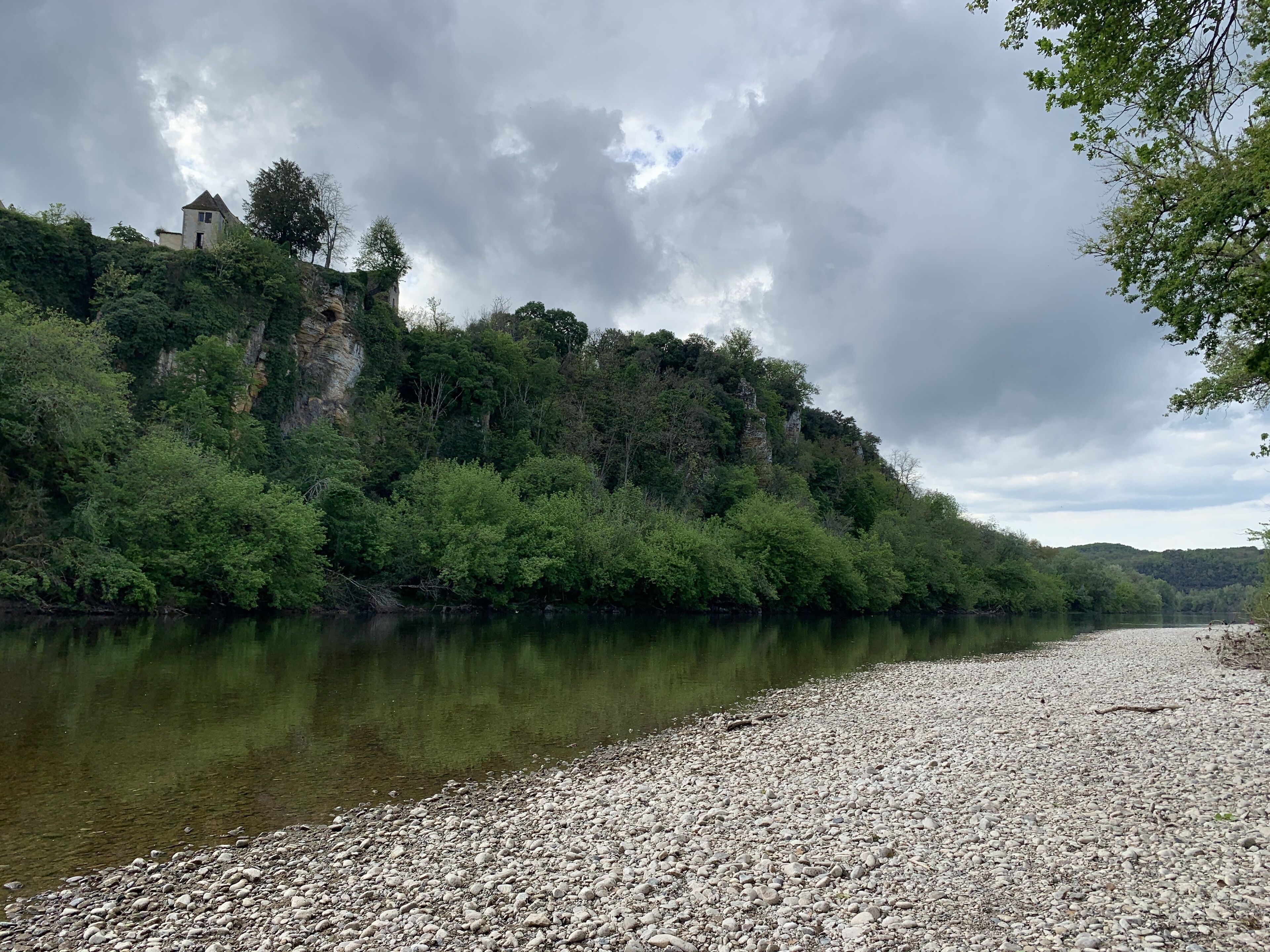A riverbank with a rocky shore, dense greenery, and a building on a cliff.