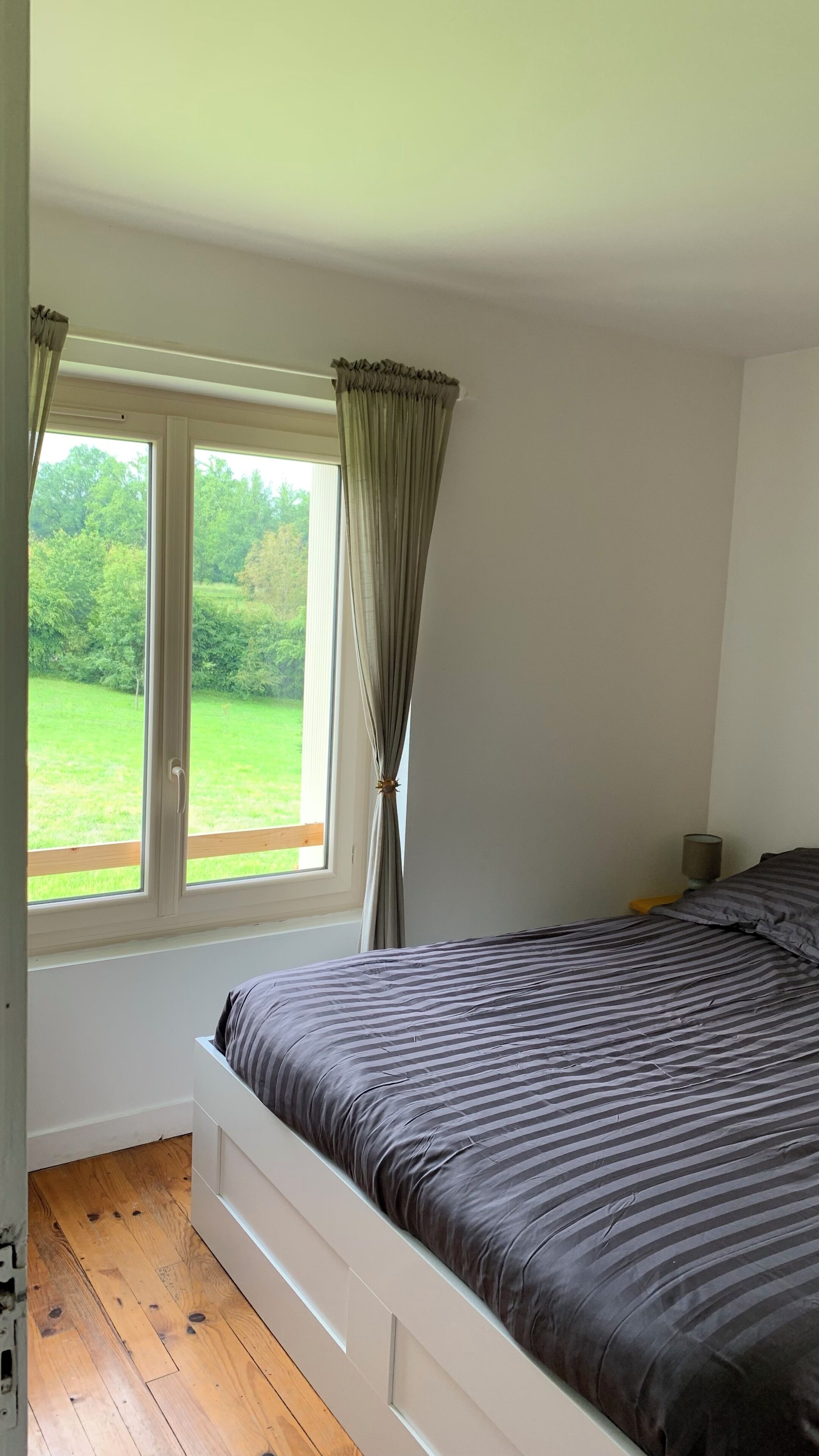 A bedroom with a large bed, a window with curtains, and a view of greenery outside.