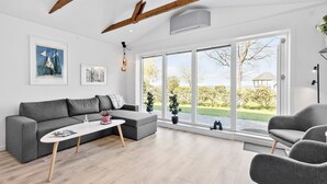 A modern living room with a grey sofa, a white coffee table, and a grey armchair. There are two potted plants, a framed picture on the wall, and a large window with a view of a garden and a gazebo.