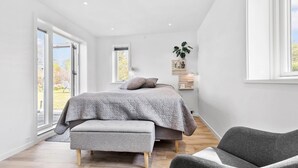 A modern bedroom with a bed, bench, and chair, large windows, and a plant on the wall.