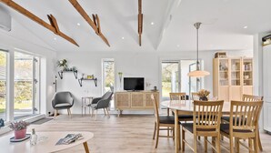 A modern living room with a dining area, wooden beams on the ceiling, a television, and a bookshelf.