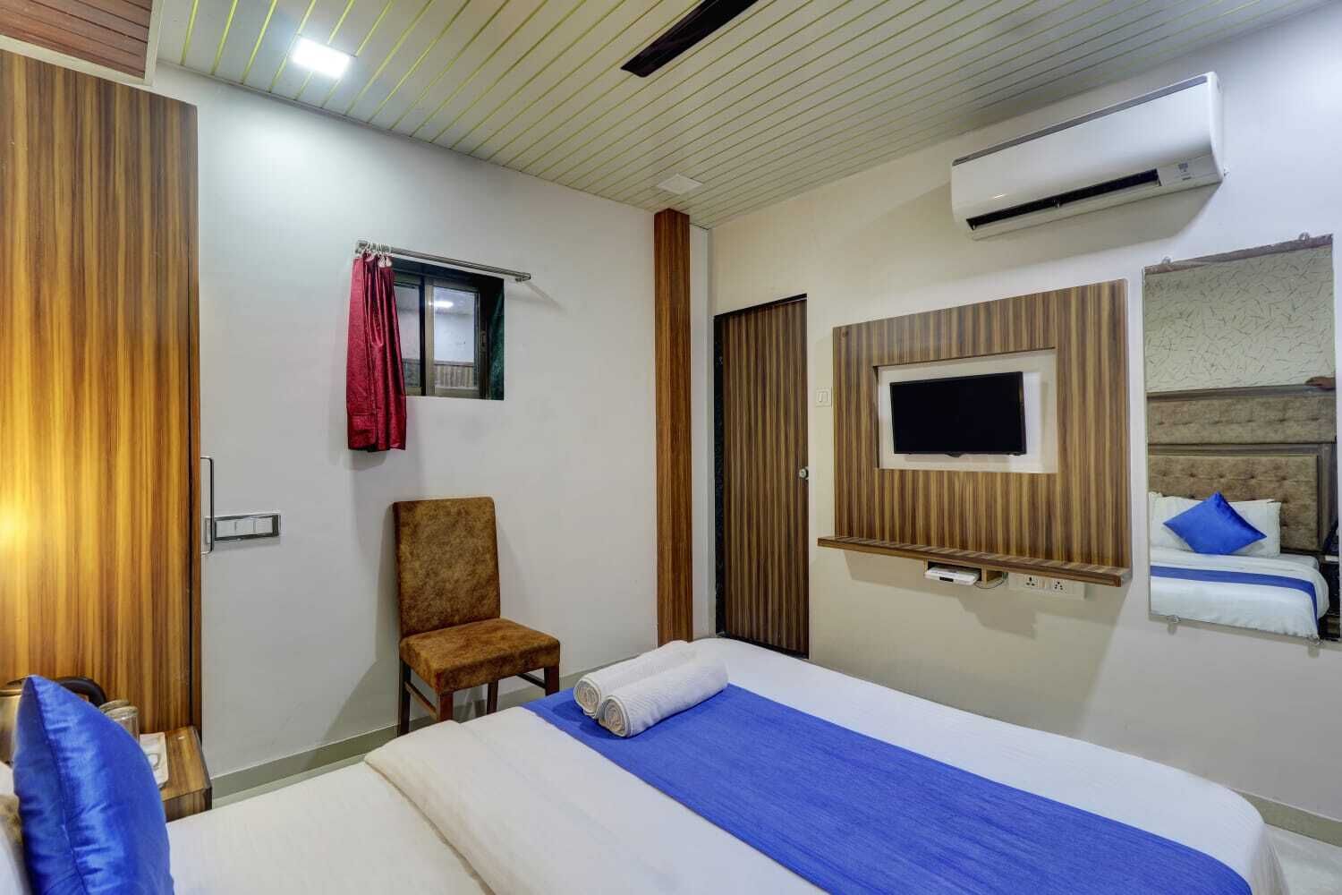 A hotel room with a bed, a chair, a TV mounted on the wall, and an air conditioning unit.