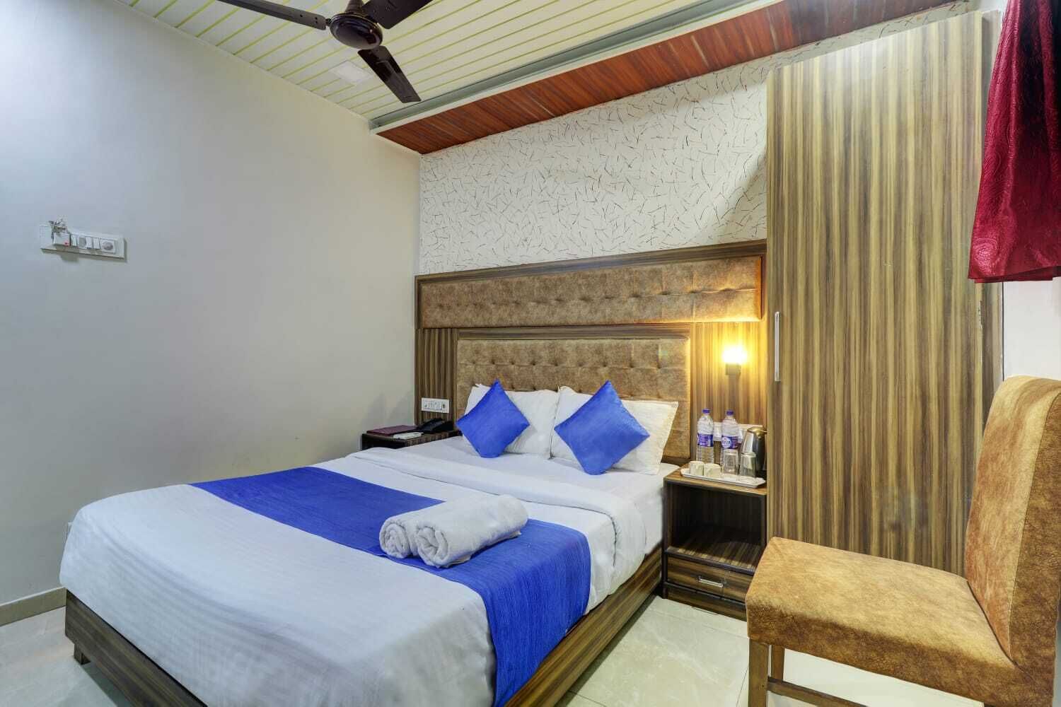 Hotelkamer met een bed, nachtkastje, stoel, plafondventilator en een wandlamp.
