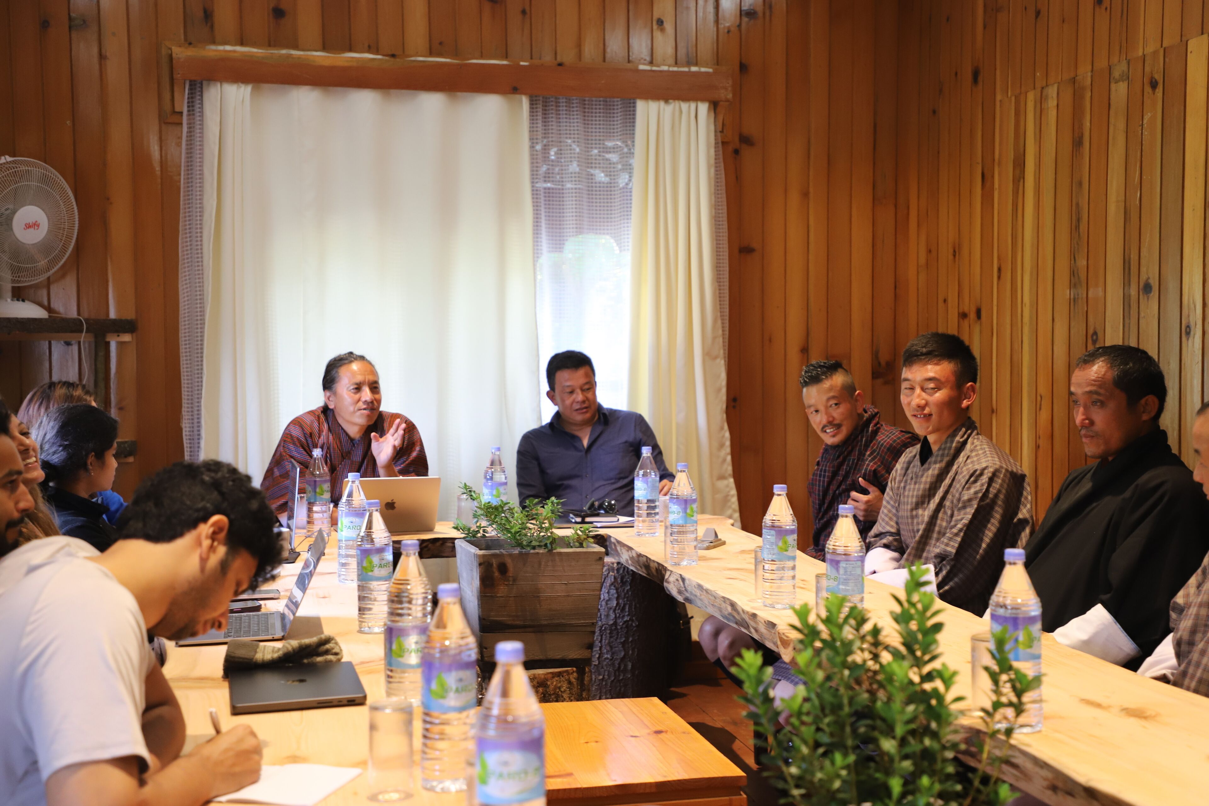 A conference room with wooden walls, a long table with laptops and water bottles, and a fan on the wall.