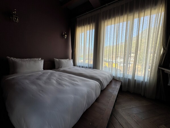 A hotel room with two beds, a wooden bench, a wall-mounted lamp, and a view of the outside through sheer curtains. - Taktsangri-La (Paro)