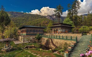 A traditional building with a stone base and a wooden upper structure, surrounded by a stone wall and a green fence, set against a mountainous backdrop. - Taktsangri-La (Paro)