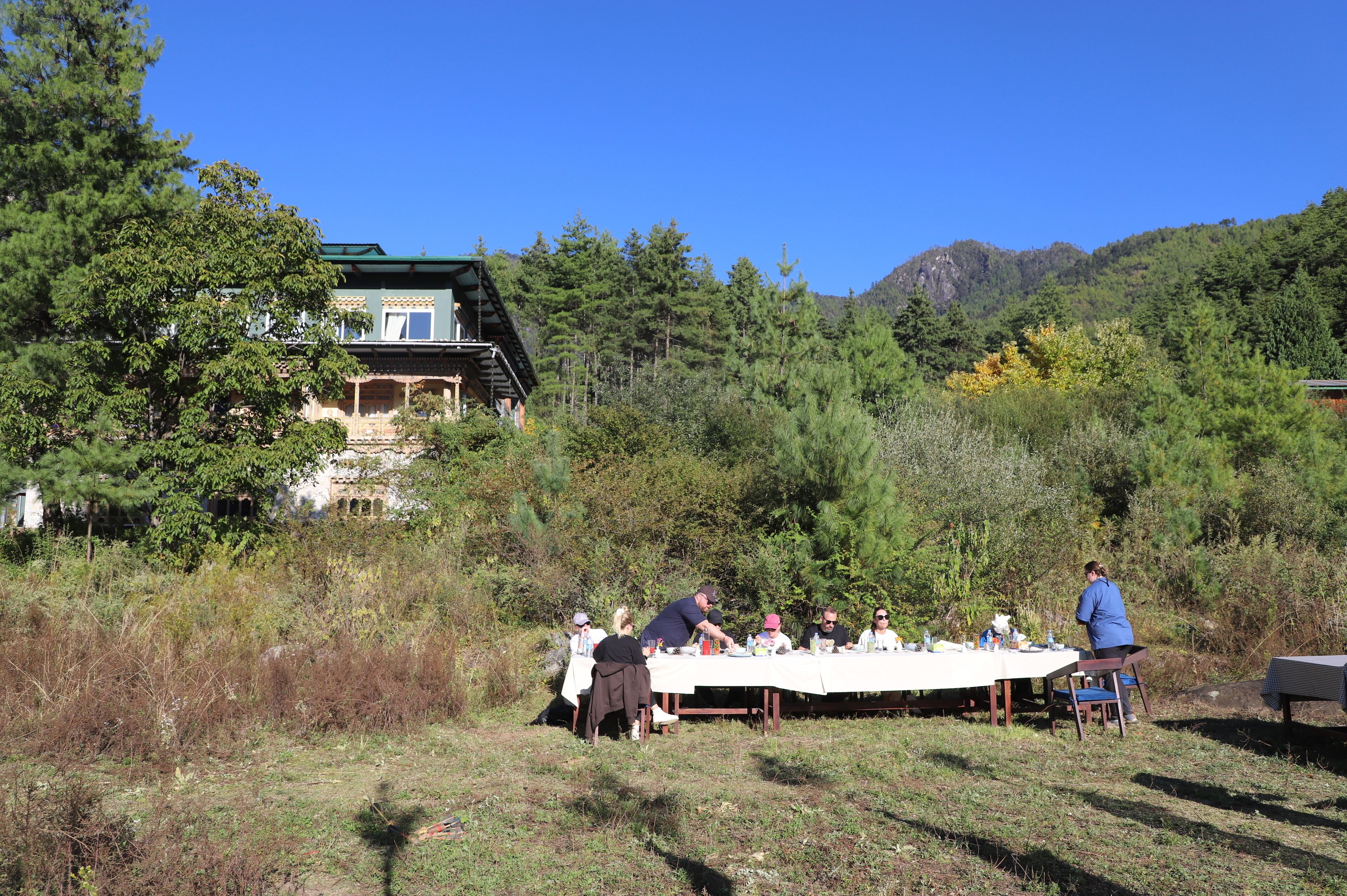 A dining table set outdoors with people seated, surrounded by trees and a building in the background.