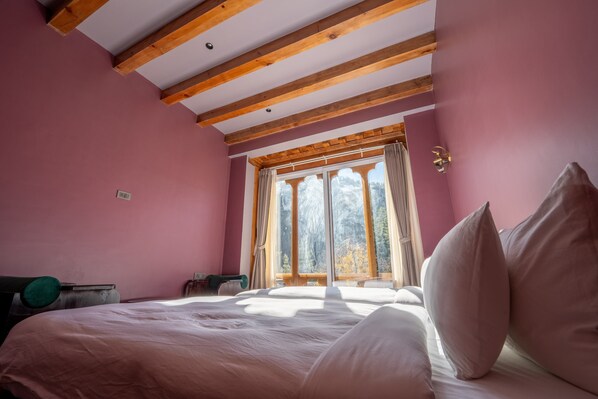 Un dormitorio con una cama grande, vigas de madera en el techo, una ventana con cortinas y vistas a árboles.