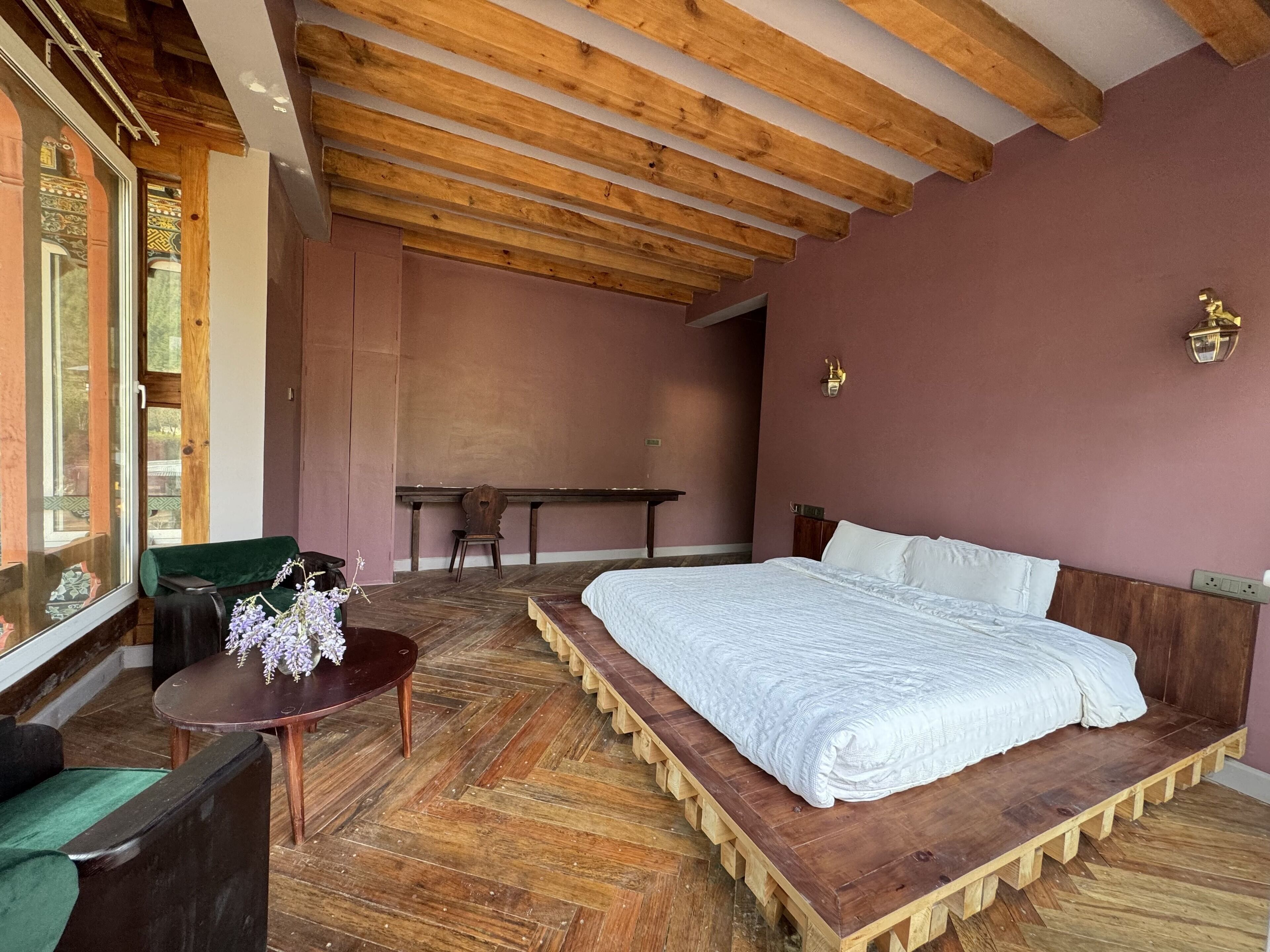 A bedroom with a large bed, wooden floor, a small table with flowers, a chair, and a bench.