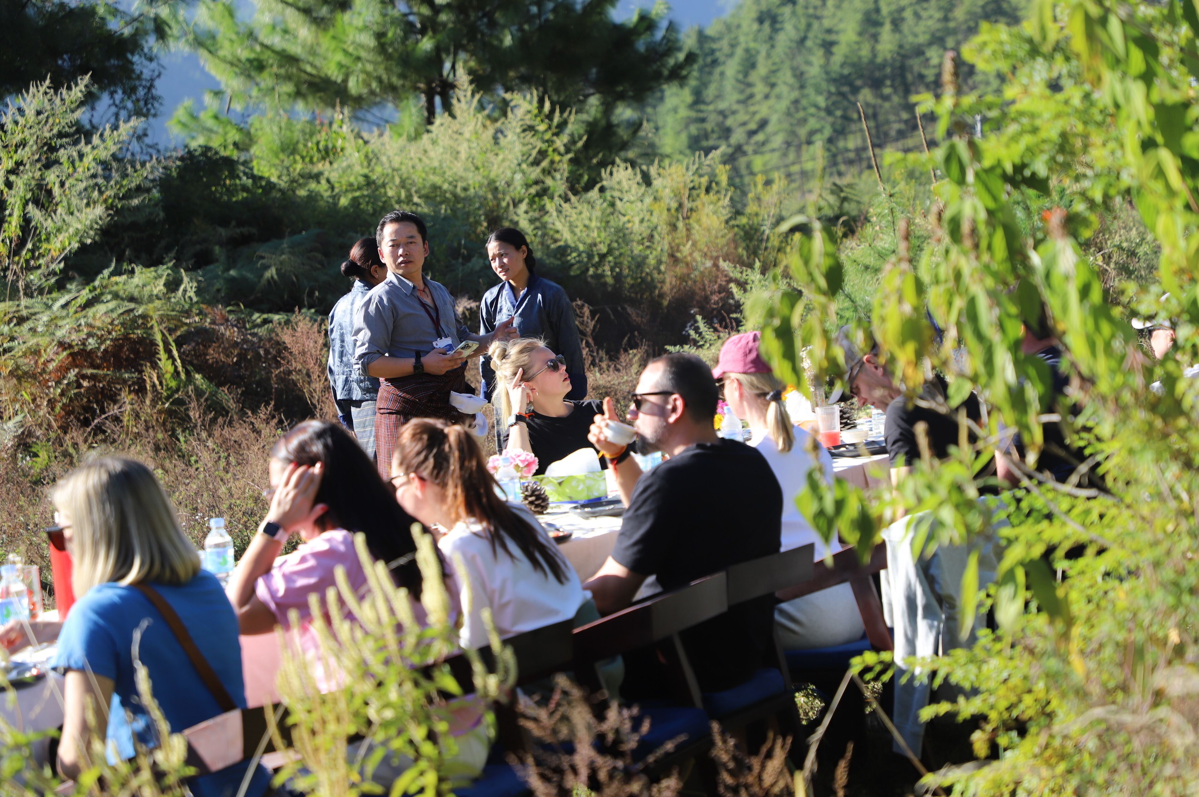 A group of people seated at an outdoor table in a natural setting with trees and greenery in the background.