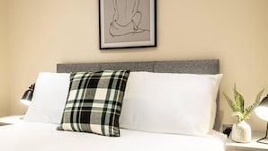 Un lit bien fait, avec du linge de lit blanc, une tête de lit grise et un coussin à carreaux verts. Un dessin linéaire encadré est accroché au mur au-dessus du lit.