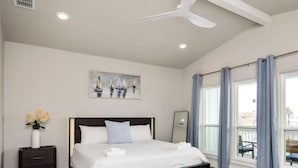 Une chambre à coucher moderne avec un lit en bois foncé, du linge de lit blanc, une table de chevet ornée d’un vase de fleurs, un ventilateur de plafond et une grande fenêtre avec des rideaux.