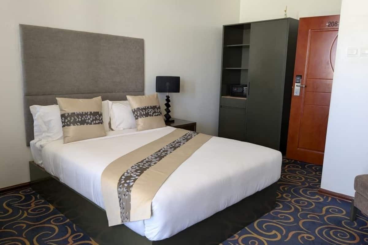 Habitación de hotel con una cama grande, una mesita de noche con lámpara, un armario y una puerta con número.