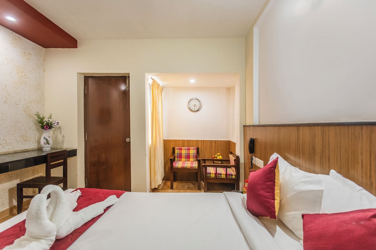 A hotel room with a bed, a desk, a chair, a small table with fruit, and a clock on the wall.