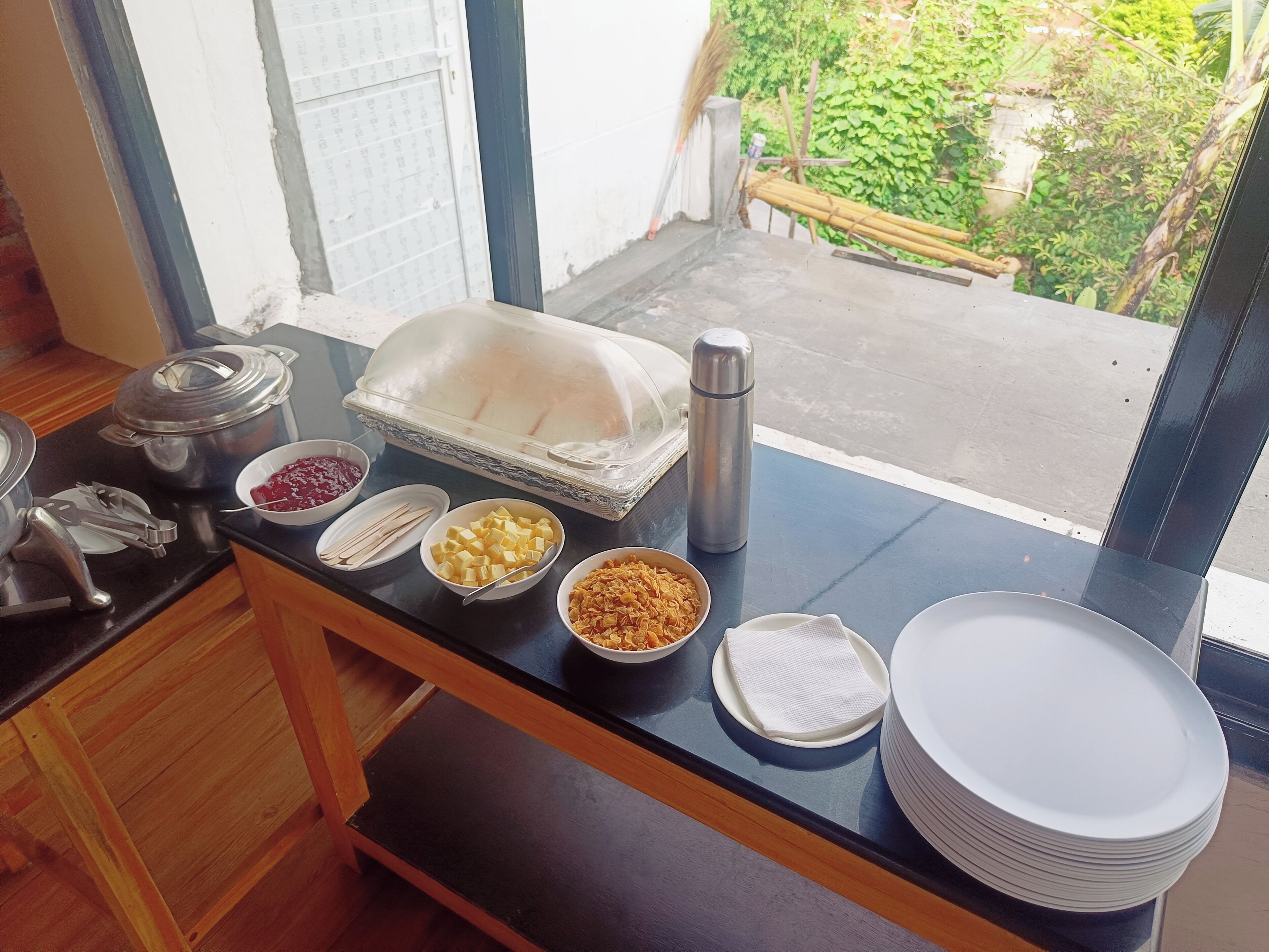 Un buffet garni de divers plats et un thermos posés sur une table près d’une fenêtre.