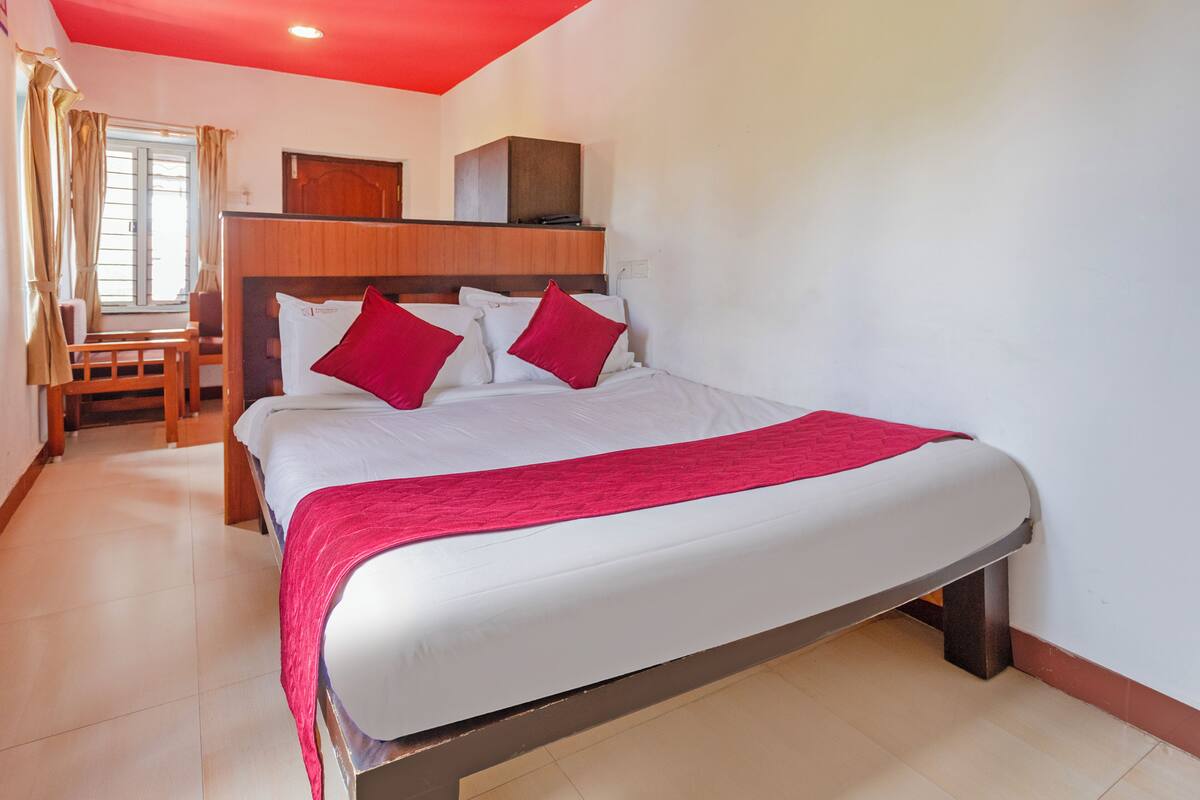 A hotel room with a bed, a wooden headboard, a red blanket, a chair, and a window with curtains.