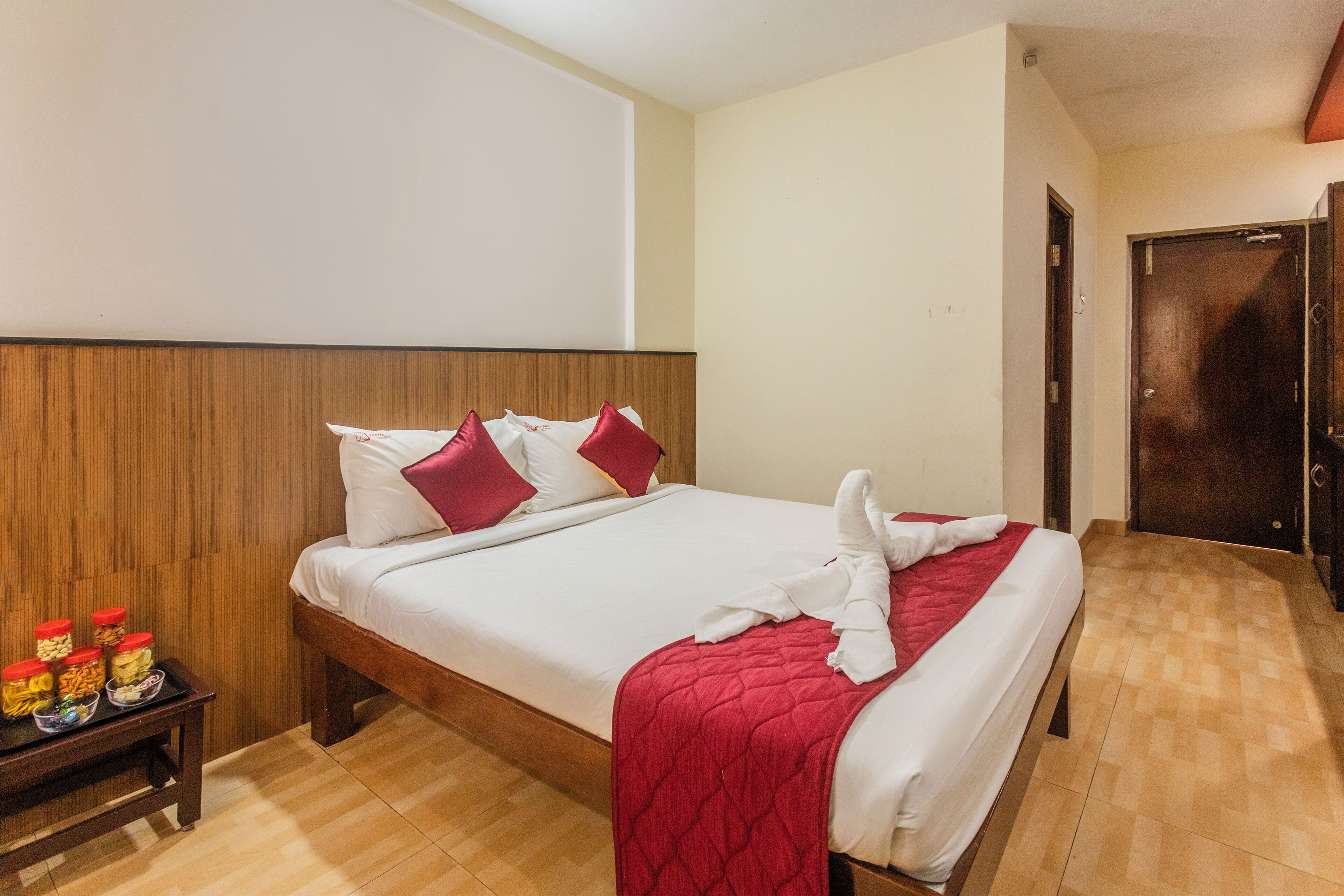 Een hotelkamer met een houten hoofdeinde, een bed met witte lakens en rode accenten, een tafeltje met etenswaren en een houten vloer.
