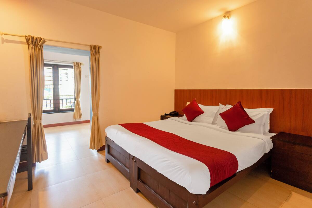 A hotel room with a large bed, wooden headboard, and a view through a window with curtains.