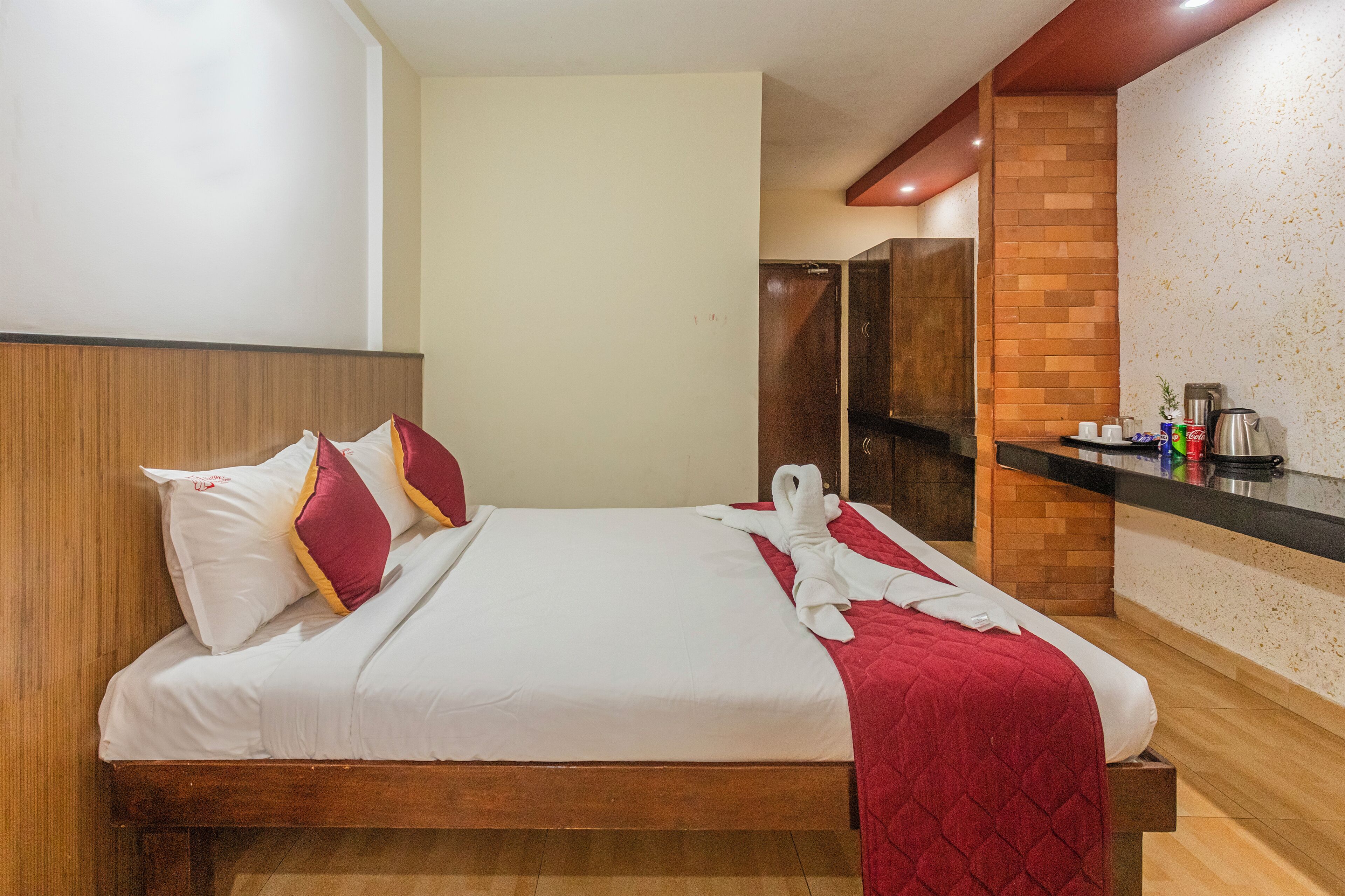 Une chambre d’hôtel avec un lit, une tête de lit en bois, un plaid rouge, une serviette, une armoire en bois et une machine à café.
