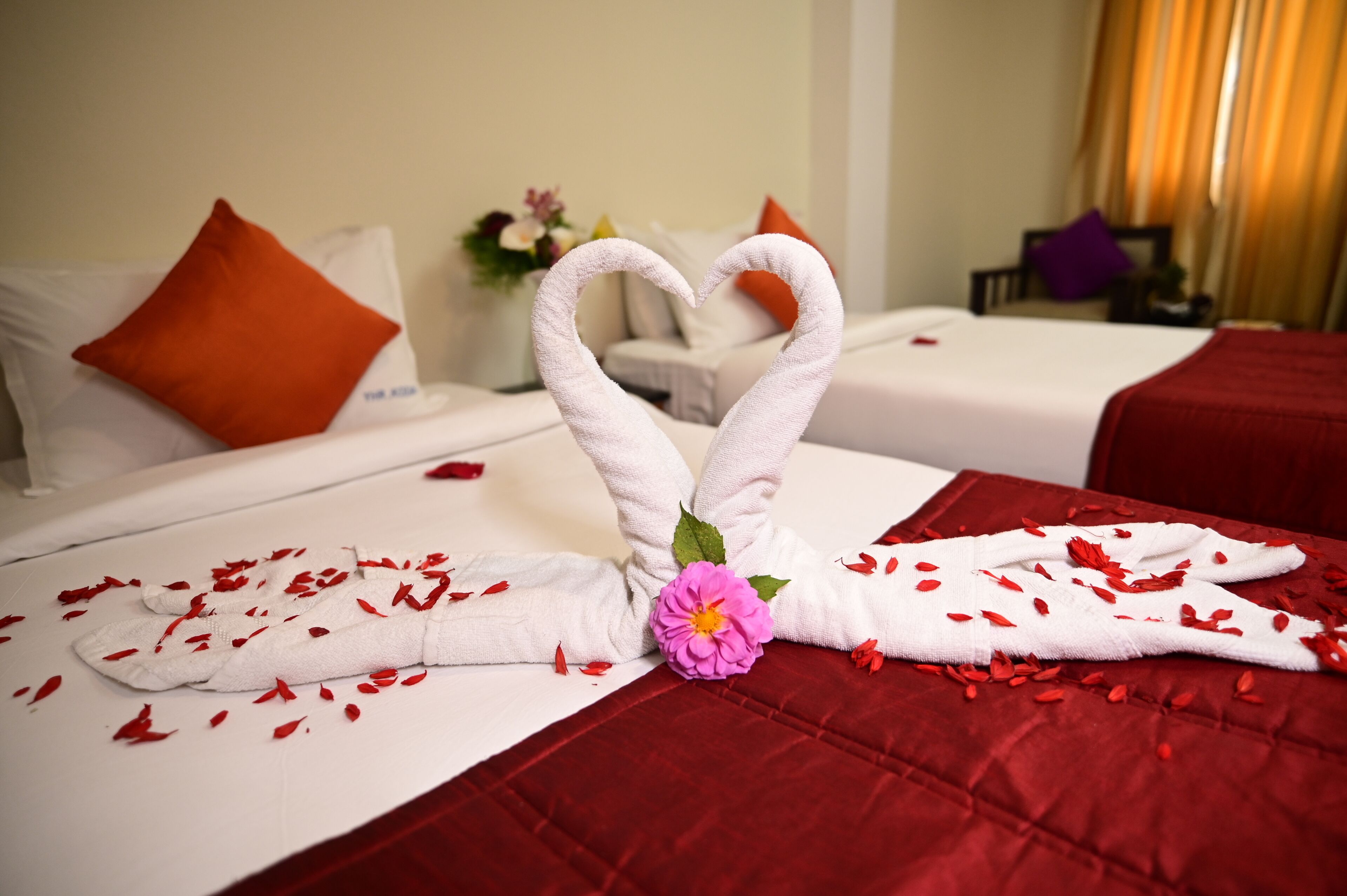 A hotel room with two beds, each with a heart-shaped towel arrangement and red flower petals.