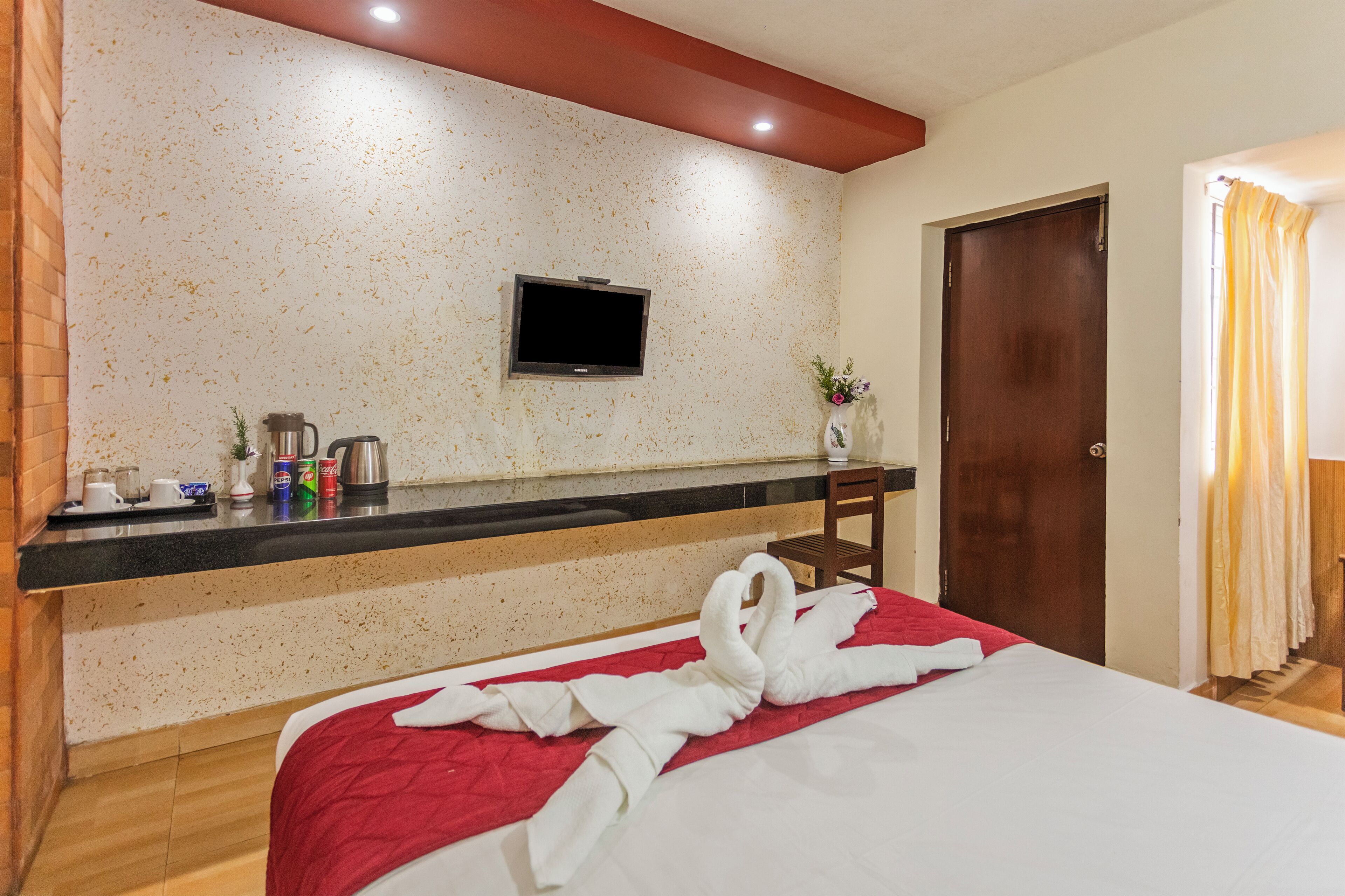 A hotel room with a bed, a TV mounted on the wall, a wooden floor, a red and white bedspread, a wooden chair, and a small table with a coffee maker and other items.
