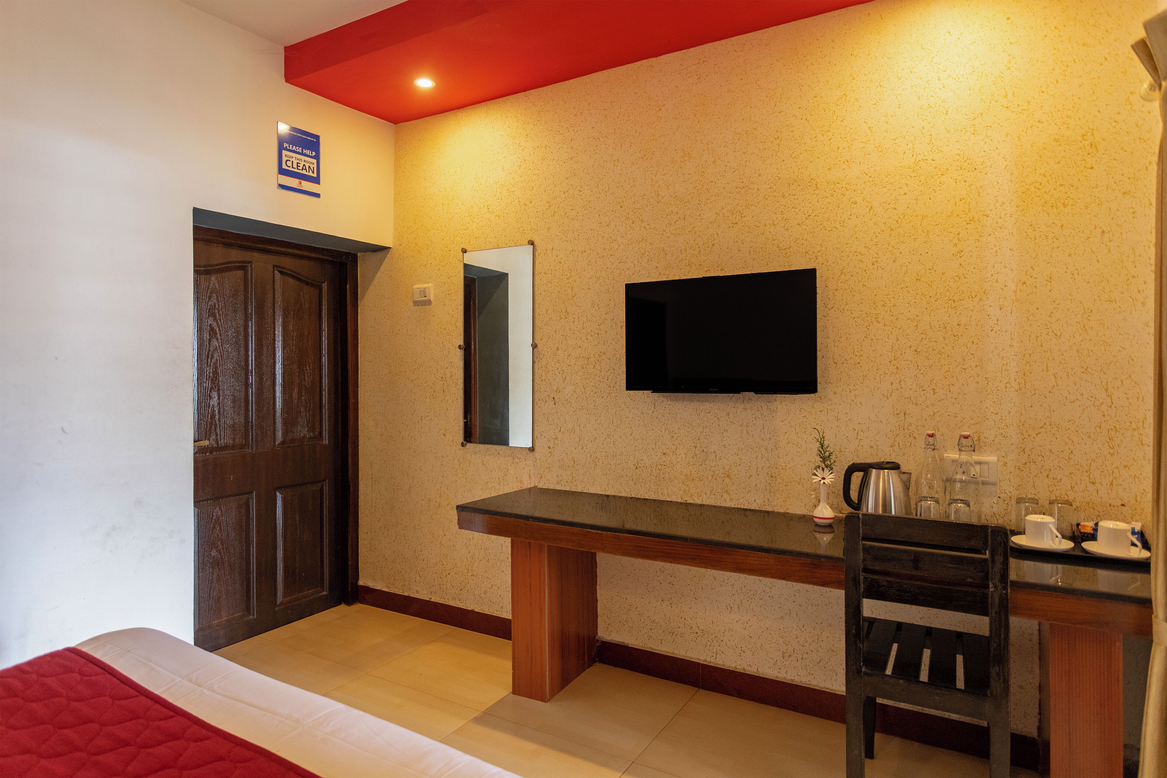 A hotel room with a wooden door, a bench, a TV mounted on the wall, and a small table with a kettle and cups.