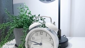 Un réveil blanc avec un socle noir, posé sur une surface blanche, à côté d’une plante en pot.