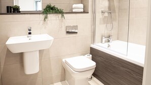 A modern bathroom with a white sink, toilet, and bathtub. There is a mirror above the sink and a showerhead above the bathtub. - One Super King Bedroom Kelham Island by Belvilla (Sheffield)