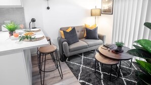 A modern living room with a grey sofa, two side tables, a round coffee table, a dining area with a white table, and a clock on the wall. - One Super King Bedroom Kelham Island by Belvilla (Sheffield)