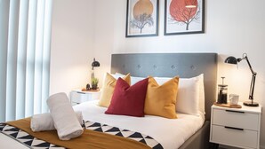 A neatly made bed with a geometric-patterned bedspread, a set of towels, and a bedside table with a lamp and a small plant. - One Super King Bedroom Kelham Island by Belvilla (Sheffield)