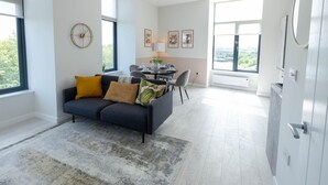 A modern living room with a dark sofa, a dining table, and a view through large windows. - Two-bedroom by Leeds Art Gallery by Belvilla (Leeds)