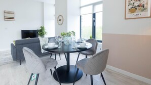 A modern dining area with a round table set for four, surrounded by grey upholstered chairs. A television is mounted on the wall to the left, and a large window provides natural light. - Two-bedroom by Leeds Art Gallery by Belvilla (Leeds)