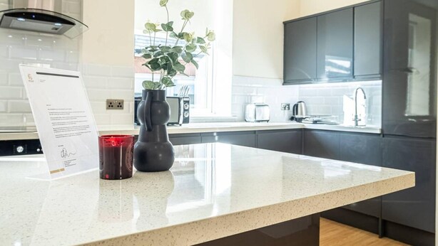 A modern kitchen with a white countertop, dark cabinets, a window, and a vase with green plants. - City Chic Christchurch 2BR Apartments by Sojo Stay (Ipswich)