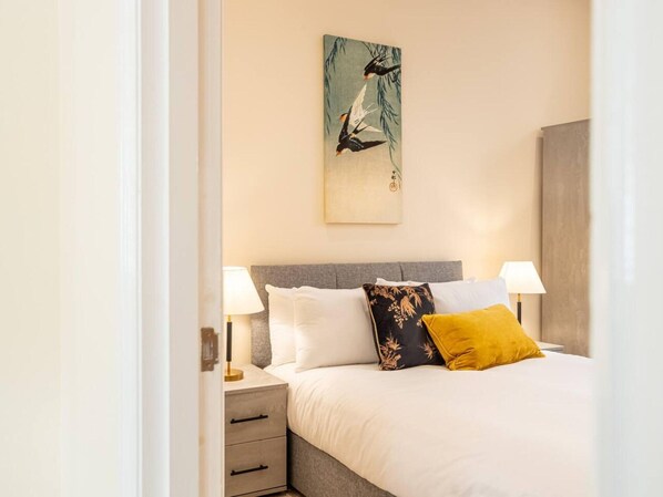 Une chambre à coucher bien rangée, avec un lit, deux lampes de chevet, une table de chevet dotée de tiroirs et un tableau accroché au mur.