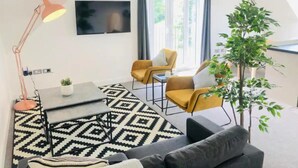 A modern living room with a sofa, two armchairs, a coffee table, and a TV. There is a patterned rug, a floor lamp, and a potted plant.