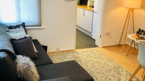 Un salon moderne avec un canapé foncé, un tapis à motifs et une petite table avec une lampe. On aperçoit une cuisine à l’arrière-plan, avec des meubles blancs et un four à micro-ondes.